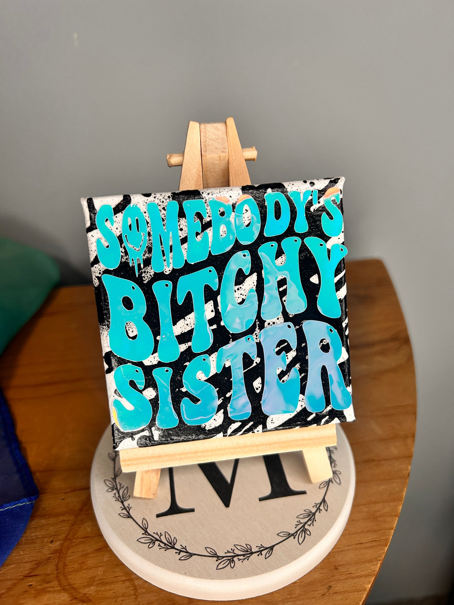 Closer view of 3 inch by 3 inch painted canvas with miniature easel, canvas is painted with black lines and over top of that is holographic permanent vinyl that says “somebody’s bitchy sister” the “o” in somebody is a slime smiley face