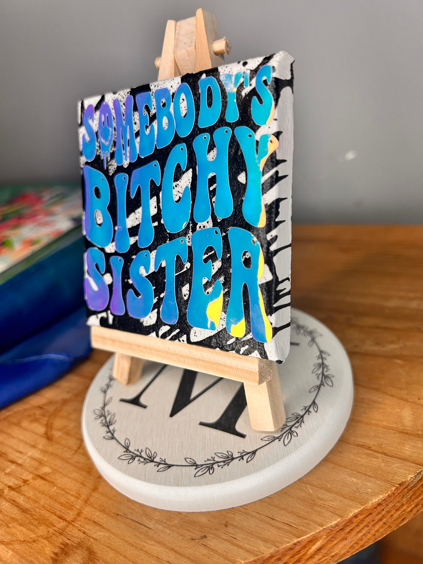 Side view of 3 inch by 3 inch painted canvas with miniature easel, canvas is painted with black lines and over top of that is holographic permanent vinyl that says “somebody’s bitchy sister” the “o” in somebody is a slime smiley face
