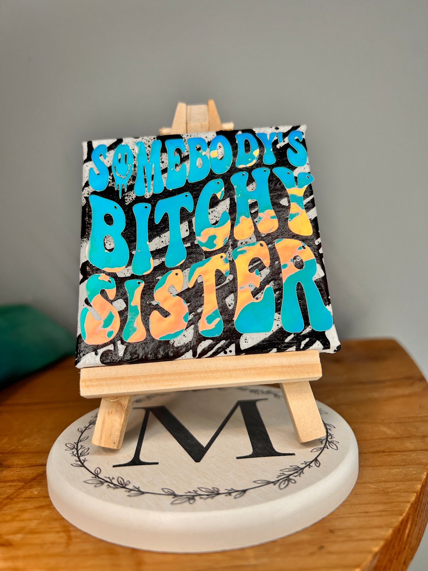 View from the bottom of 3 inch by 3 inch painted canvas with miniature easel, canvas is painted with black lines and over top of that is holographic permanent vinyl that says “somebody’s bitchy sister” the “o” in somebody is a slime smiley face