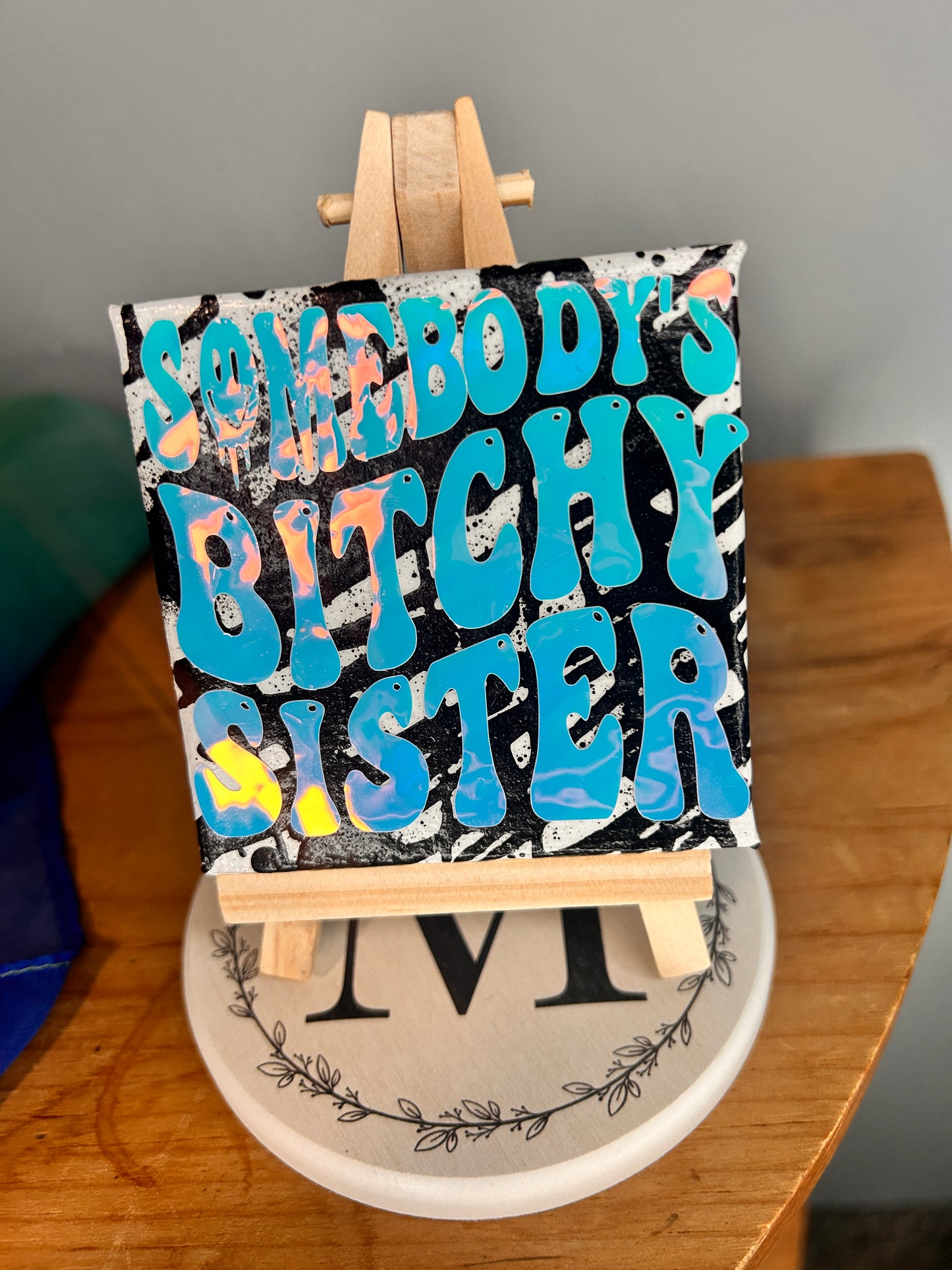 Photo showing the holographic effects of 3 inch by 3 inch painted canvas with miniature easel, canvas is painted with black lines and over top of that is holographic permanent vinyl that says “somebody’s bitchy sister” the “o” in somebody is a slime smiley face