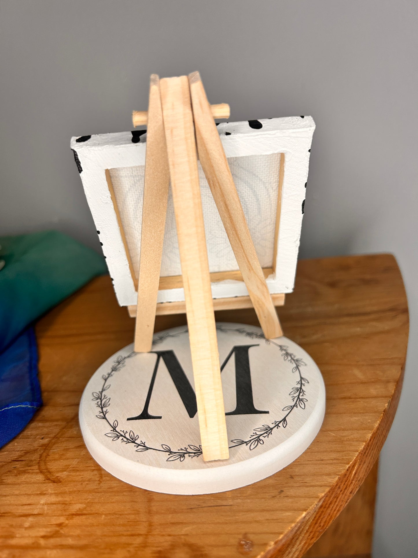 Back view of the miniature easel holding the 3 inch by 3 inch painted canvas with miniature easel, canvas is painted with black lines and over top of that is holographic permanent vinyl that says “somebody’s bitchy sister” the “o” in somebody is a slime smiley face