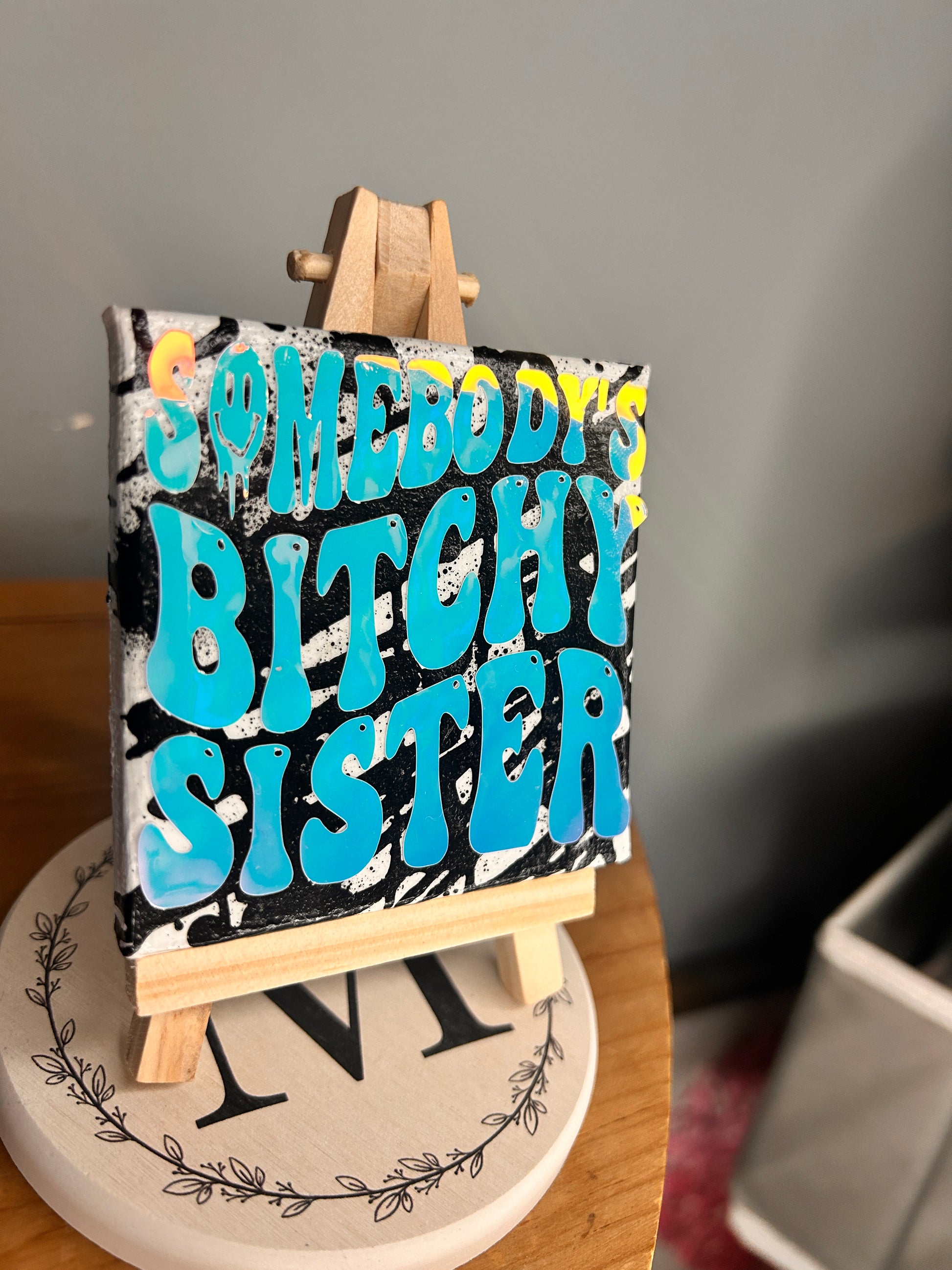 3 inch by 3 inch painted canvas with miniature easel, canvas is painted with black lines and over top of that is holographic permanent vinyl that says “somebody’s bitchy sister” the “o” in somebody is a slime smiley face 